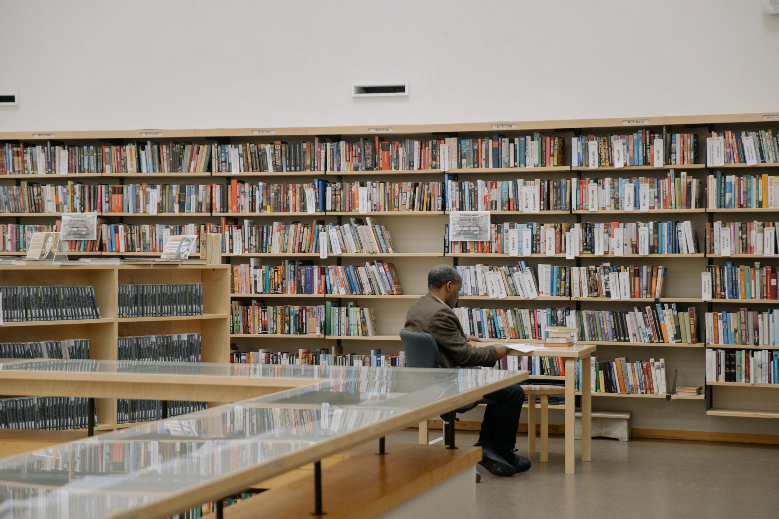 Man sitting at a table reading in a well-lit modern library surrounded by bookshelves.